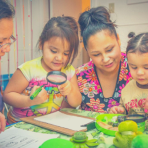 Two women with dark hair and darker skin smile while looking at a clipboard with two young children. Both children have dark skin and dark hair and are looking at the clipboard through a magnifying glass.